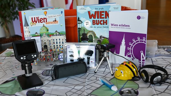 Inhalte der verschiedenen Digiboxen sind auf einem Tisch aufgestellt: Wien-Matte, Bee-Bot, Audioaufnahmegerät, Bücher, ein digitales Mikroskop, ein "Wien erleben"-Booklet und einiges mehr.